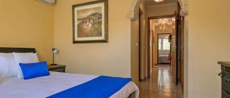 A bedroom with a bed, a nightstand, a lamp, a painting, and a hallway leading to another room.