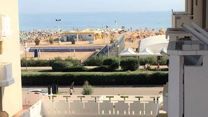 A view of a beach with umbrellas and people, a road with a cyclist, and a building with a balcony. - Holiday apartment on the beach (Bibione)