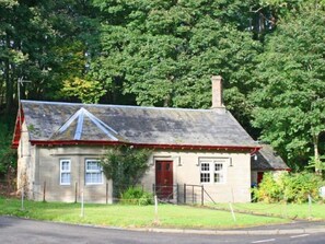 Une petite maison en pierre, de plain-pied, avec une porte rouge, une cheminée et une cour clôturée.