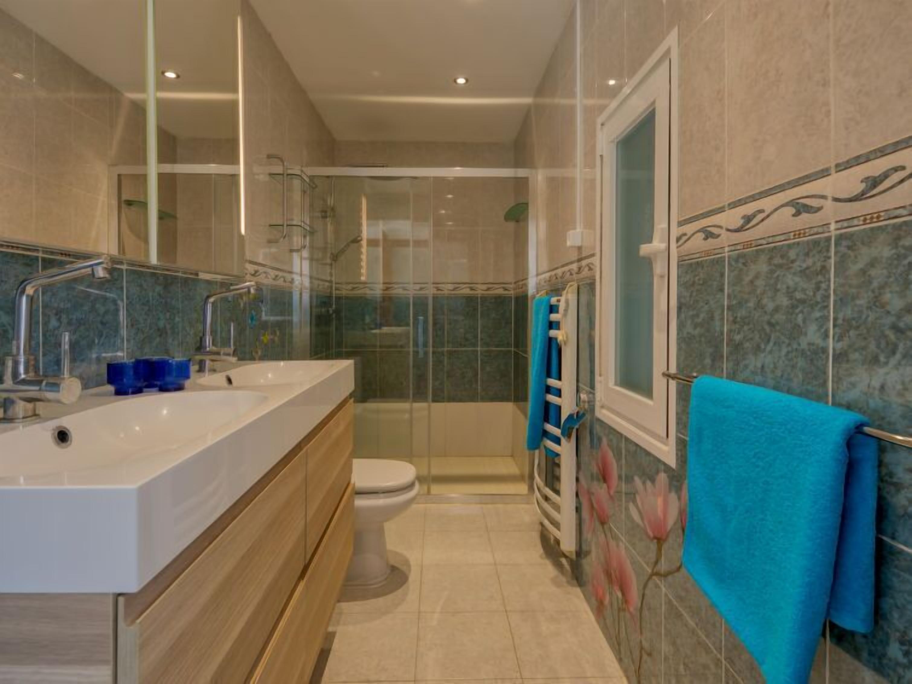 A modern bathroom with a dual sink vanity, a toilet, a glass-enclosed shower, and a towel rack with blue towels.