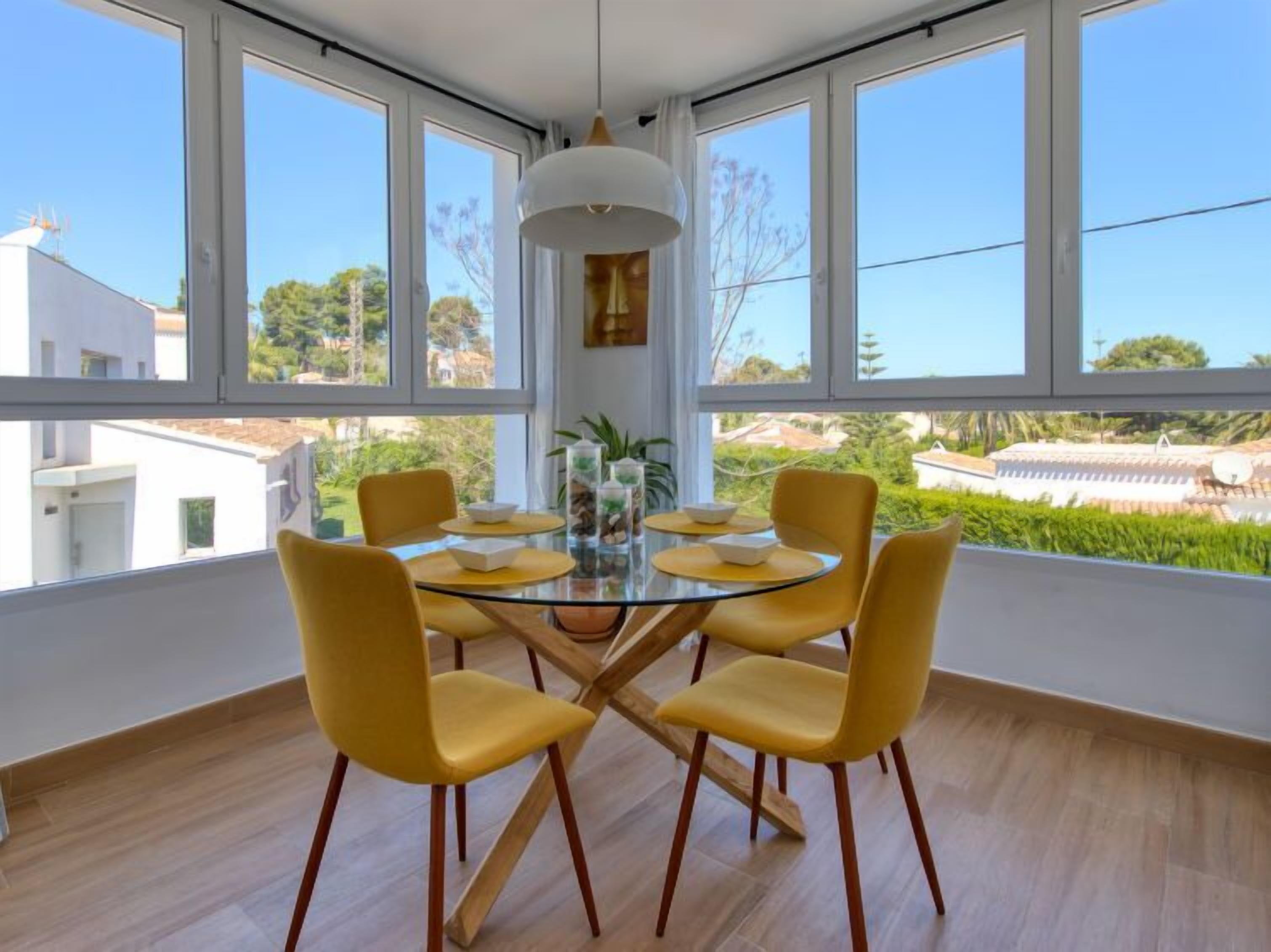A dining area with a glass table and yellow chairs, large windows, and a view of greenery and buildings.