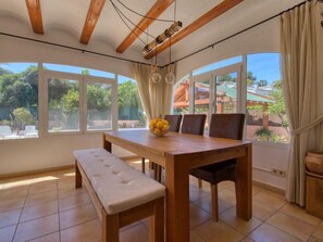 A dining area with a wooden table, bench, and chairs, large windows with curtains, and a view of greenery outside.