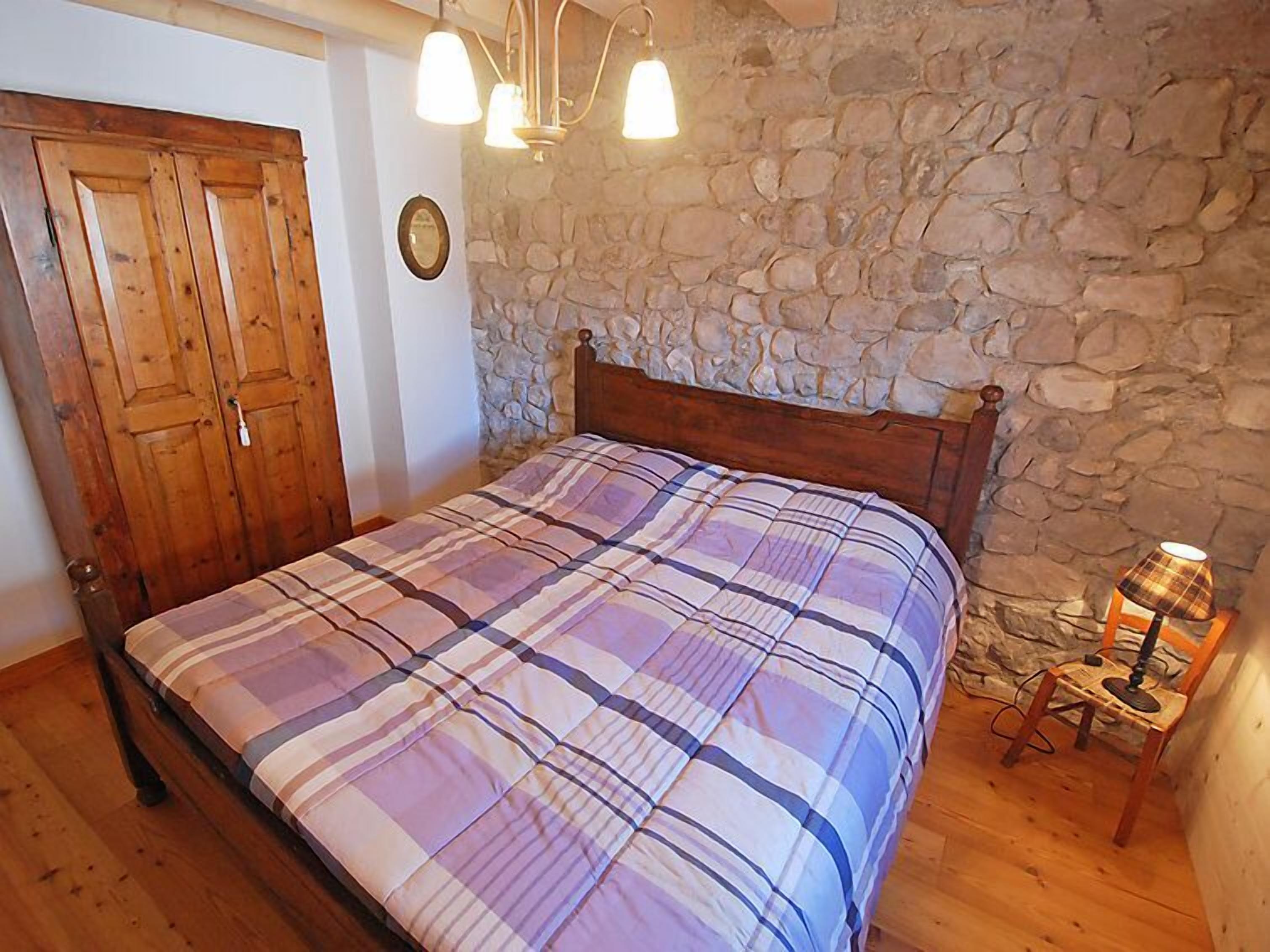 Quarto com cama de madeira, parede de pedra e uma pequena mesa com abajur.