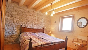 Une chambre avec un mur en pierre, des poutres en bois au plafond, un lit recouvert d’une couverture à carreaux, une chaise en bois et une fenêtre avec un miroir.