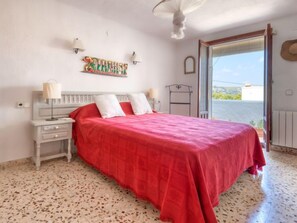 Un dormitorio con colcha roja, cabecero blanco y vistas al mar a través de una puerta abierta.