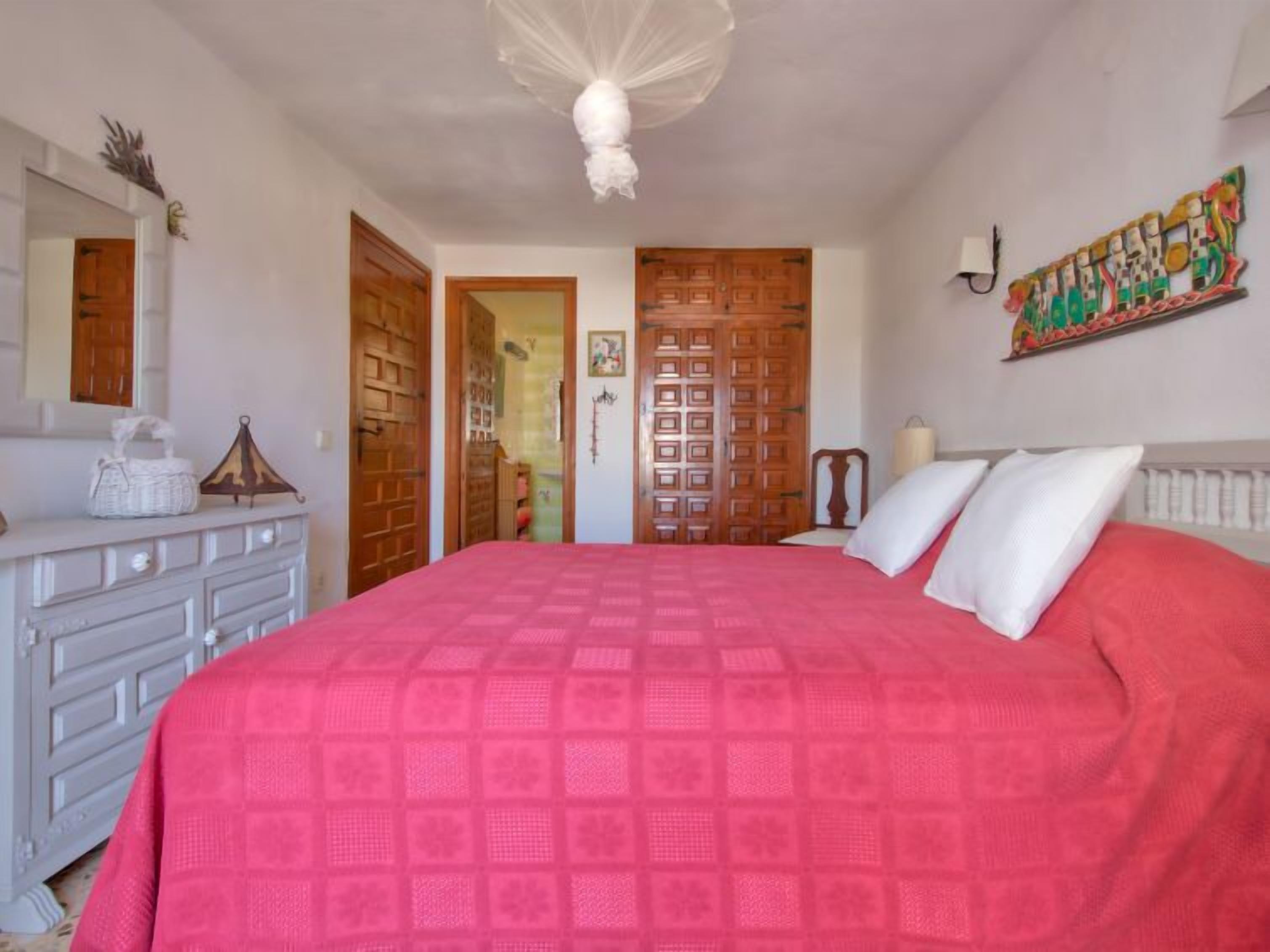 A bedroom with a large bed, wooden doors, a dresser, and a wall-mounted lamp.