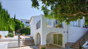 A white building with a stone staircase, a small patio with a chair and table, and a tree providing shade. - Florencia by Interhome (Jávea)