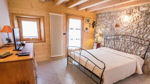 A bedroom with a bed, wooden walls, a stone wall, a window, a radiator, a desk with a lamp, and a television. - Albergo Diffuso - Cjasa Ustin by Interhome (Barcis Lake)