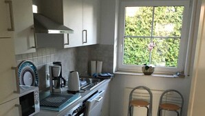 Een kleine keuken met witte kastjes, een fornuis, een koffiezetapparaat en een raam met uitzicht op groen.