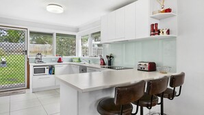 Une cuisine moderne avec des meubles blancs, un grand îlot central doté de tabourets, un grille-pain et une vue sur le jardin à travers une porte vitrée.