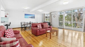 Un salon spacieux avec un canapé en cuir rouge, une table basse en bois et un coin repas comprenant une table et des chaises blanches. Une grande fenêtre offre une vue sur les arbres et un immeuble.
