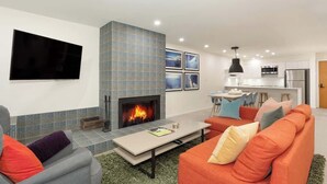 A modern living room with a fireplace, a flat-screen TV, and a kitchen in the background. - Cozy condo w/ hot tub & fireplace near lifts (Whistler)