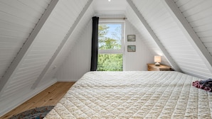 Quarto com teto inclinado, cama com cobertura acolchoada, mesa de cabeceira com abajur e janela com vista para árvores.