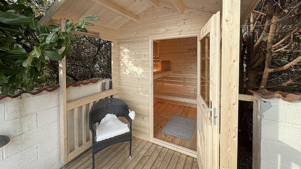 Uma sauna de madeira com porta de vidro, uma cadeira de vime com almofada branca e um tapete pequeno no chão.
