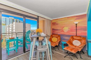 A balcony with turquoise chairs, a table with a vase of flowers, and a sunset mural on the wall.