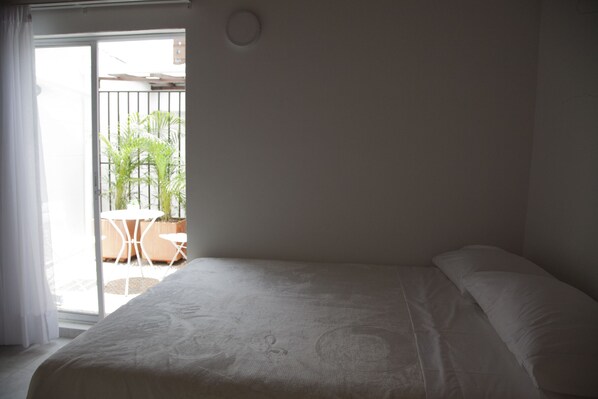 A bedroom with a bed, a sliding glass door leading to a balcony with a table and chairs, and a potted plant.