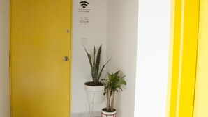 Een gang met een gele deur, een kleine plant in een pot op een rode kruk, en een Wi-Fi symbool aan de muur.
