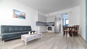Un apartamento moderno con un sofá gris, una mesa de centro, una mesa de comedor con sillas, una cocina con armarios blancos y un salpicadero azul, y un cuadro de un paisaje urbano.