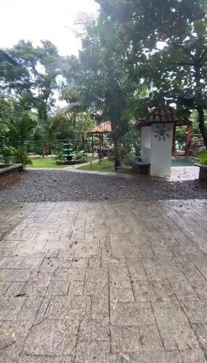 A paved outdoor area with a gazebo, surrounded by trees and a grassy lawn.