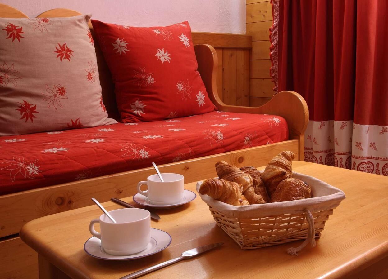 A cozy room with a wooden bed, red bedding, two white cups with spoons, and a basket of croissants on a wooden table.