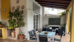 Un patio couvert comprenant une table et des chaises, des plantes en pot et un espace détente avec des coussins.