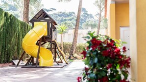 Une structure de toboggan jaune dans un espace extérieur aménagé, avec un chemin pavé, entourée de verdure et d’un bâtiment en arrière-plan.