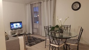 Un salon avec un téléviseur, une table à manger avec des chaises, une petite table ronde avec un vase et une horloge au mur.