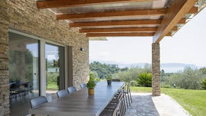 A covered outdoor dining area with a long table and chairs, stone walls, and a view of the countryside. - Villa with park, private pool and lovely lake view (Gardone Riviera)