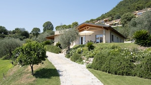 A modern house with a stone pathway leading to the entrance, surrounded by lush greenery and hills. - Villa with park, private pool and lovely lake view (Gardone Riviera)