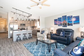 Ein modernes Wohnzimmer mit einem schwarzen Ledersofa, einem Glastisch und einem Teppich mit blauem Muster. Es gibt eine Küchenzeile mit Kühlschrank, Mikrowelle und Barhocker. Der Raum verfügt über einen Deckenventilator und Wandleuchten.