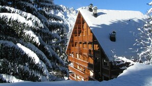A wooden cabin with a steep roof, surrounded by snow-covered trees and mountains.