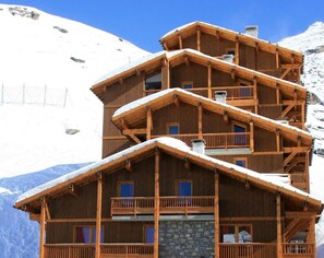 A multi-story wooden chalet with balconies, set against a snowy mountain backdrop.