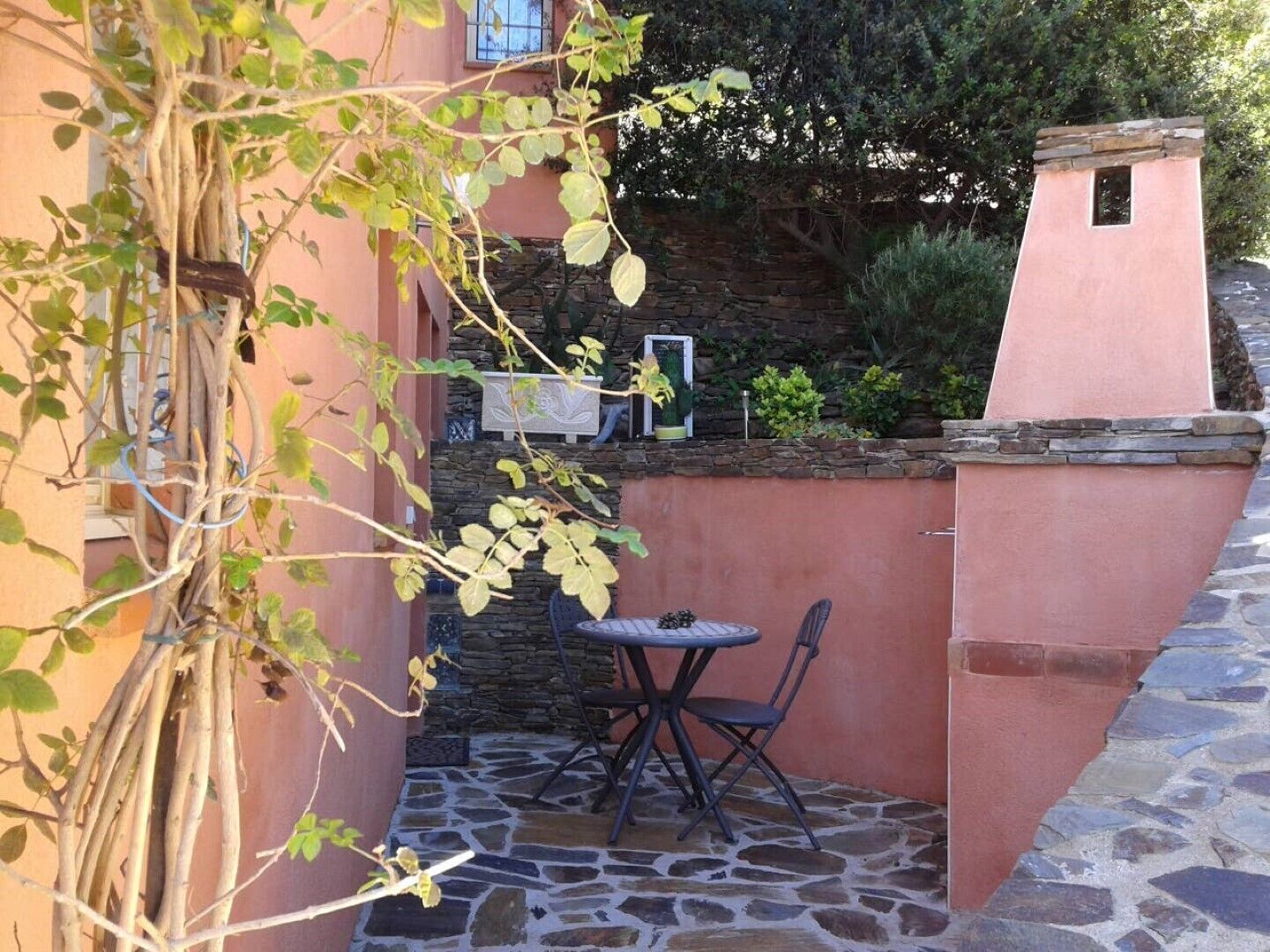Un pati de pedra amb taula i caires, una petita paret de pedra, i un edifici amb façana rosa.