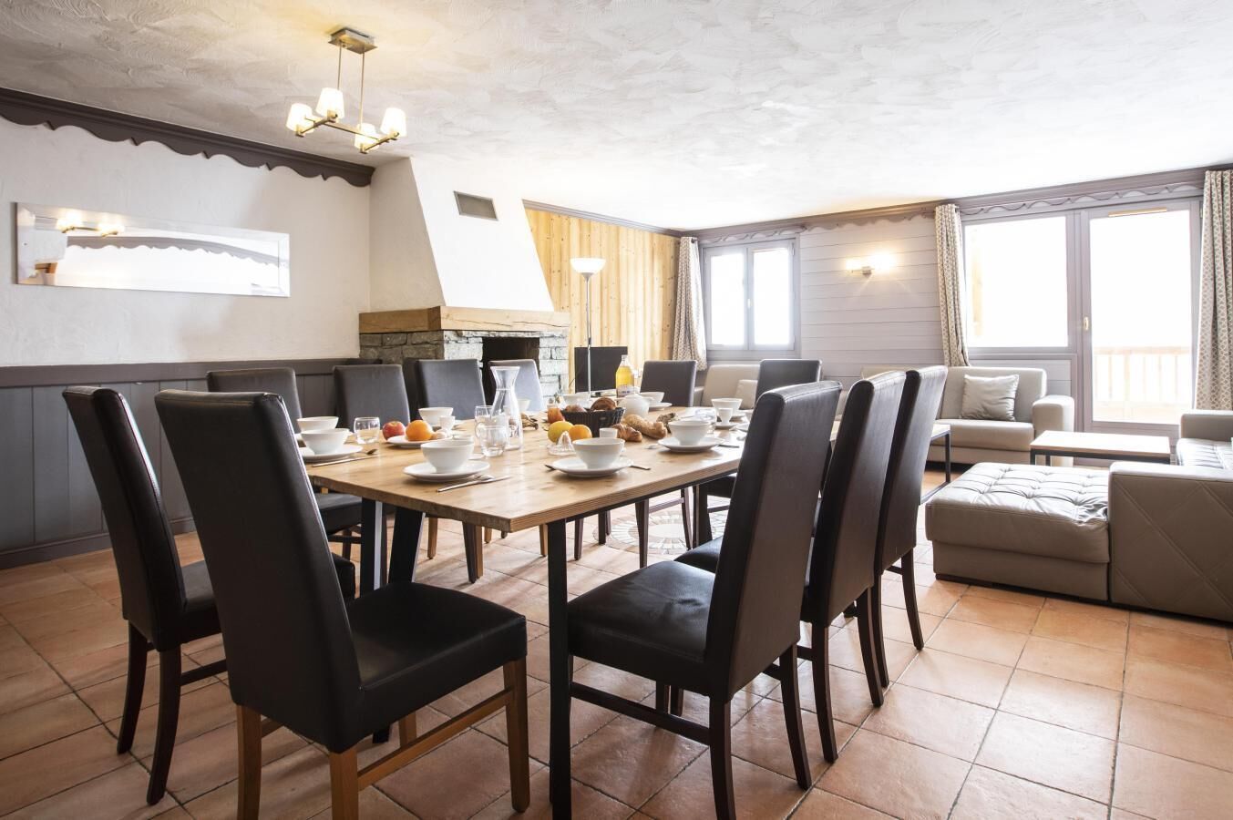 Ein Essbereich mit einem Holztisch, gedeckt für vier Personen, umgeben von schwarzen Stühlen. Im Hintergrund befindet sich eine Couch und ein Fenster mit Vorhängen.
