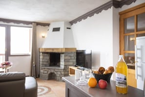 A cozy living room with a stone fireplace, a television, and a counter with bread, fruit, and a bottle of juice. - Wifi, 200m from resort center, fitness, swimming pool, sauna, balcony, fireplace or stove, tv (Val Thorens)