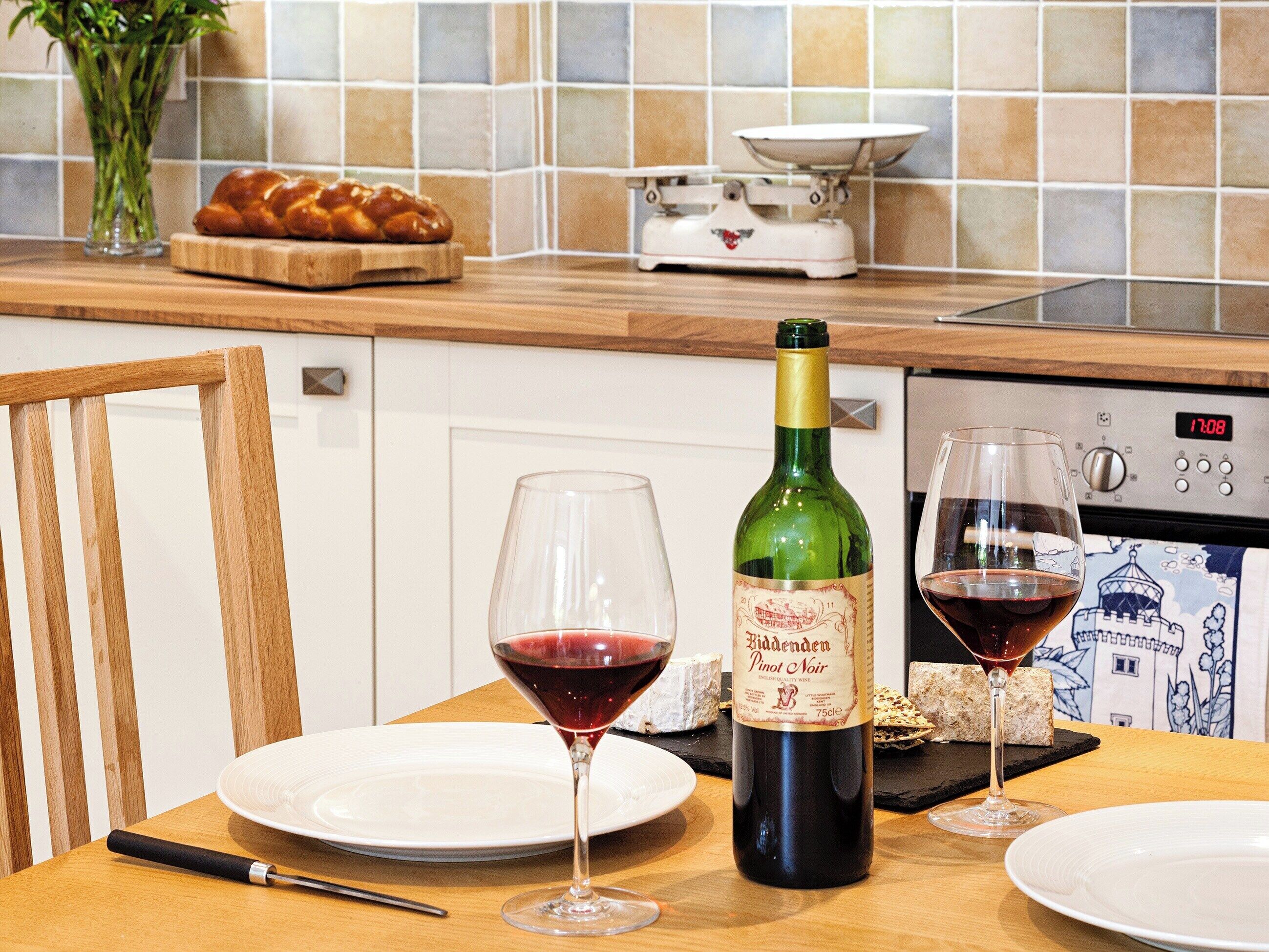 Une table à manger avec une bouteille de vin, deux verres à vin, un pain et une balance de cuisine posés sur un plan de travail.