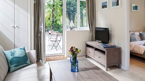 A room with a sofa, a TV on a wooden stand, a dining table with chairs, and a view of greenery through the window. - A lovely, modern apartment just 350 yards from the beach. (St Margaret&rsquo;s Bay)