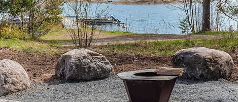 A lakeside view with a fire pit, large rocks, and a dock in the background.