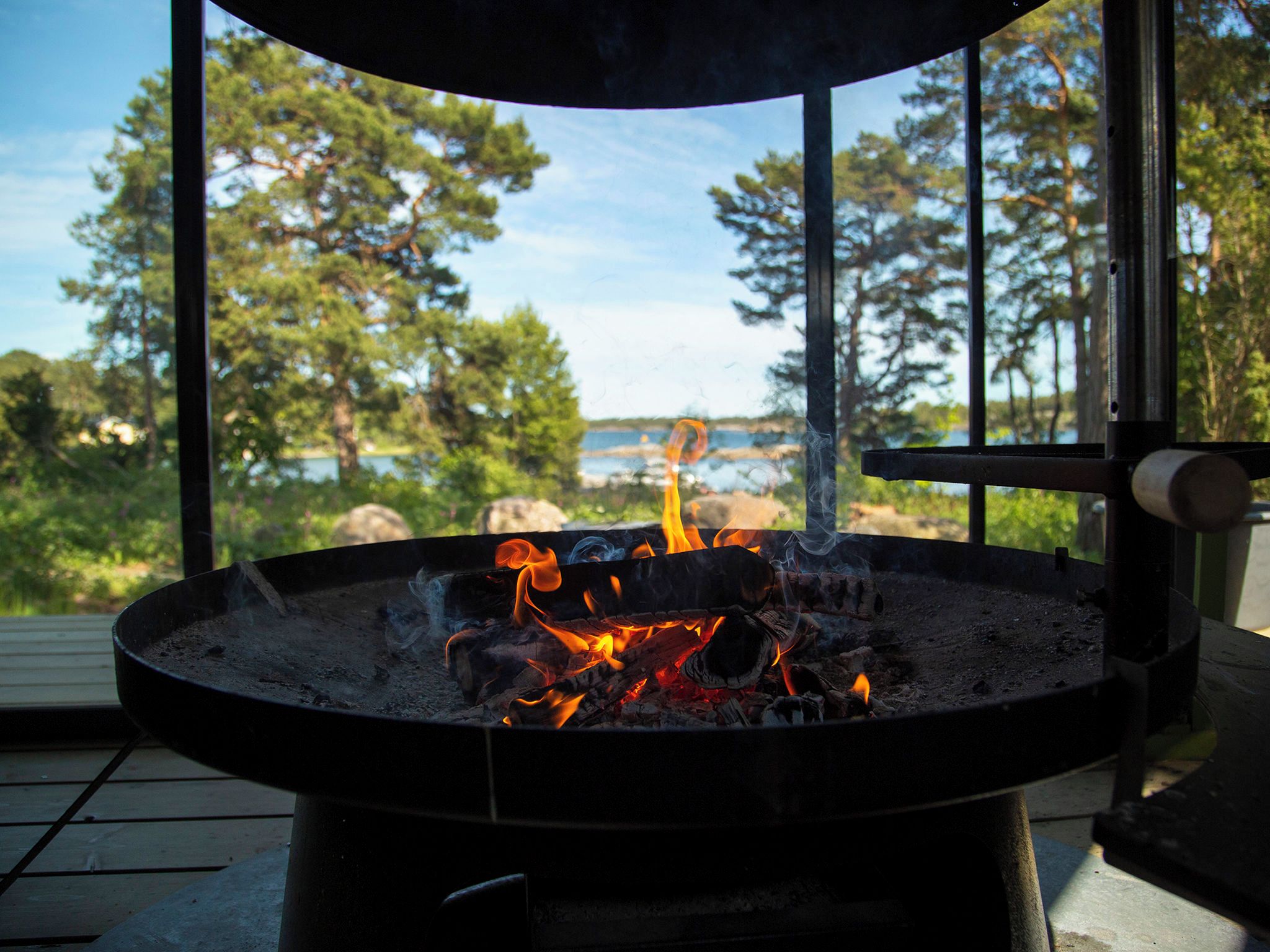A fire pit with flames, surrounded by a metal frame, set on a wooden deck with a view of trees and a body of water.