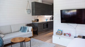 Una sala de estar moderna con un sofá, dos mesitas, un televisor de pantalla plana y una zona de cocina con armarios oscuros y una encimera blanca.