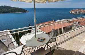 A balcony with a table, chairs, and an umbrella overlooking a coastal view with a distant island and a fortified town.
