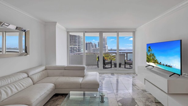 Una sala moderna con un sofá seccional color beige, una mesa de centro de vidrio y un televisor de pantalla plana sobre un mueble blanco. Amplias ventanas ofrecen vistas del horizonte de la ciudad y un balcón con mobiliario exterior.