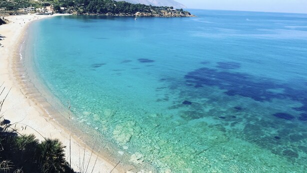 Una playa costera con aguas turquesas cristalinas, una orilla arenosa y montañas al fondo.