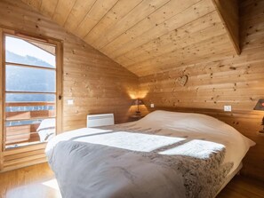 A cozy wooden cabin room with a bed, a nightstand, a lamp, and a window overlooking a snowy landscape.
