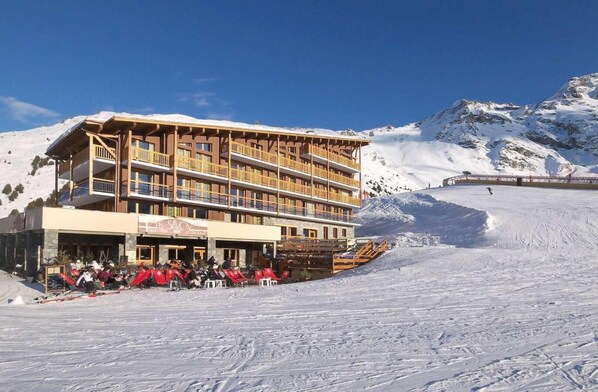 Un hôtel à plusieurs étages doté d’une terrasse, de pistes de ski et, en arrière-plan, de montagnes enneigées.