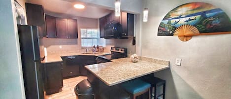 Una cocina con encimeras de granito, armarios oscuros, un refrigerador negro y un microondas. Hay dos taburetes de bar y un cubo de basura.