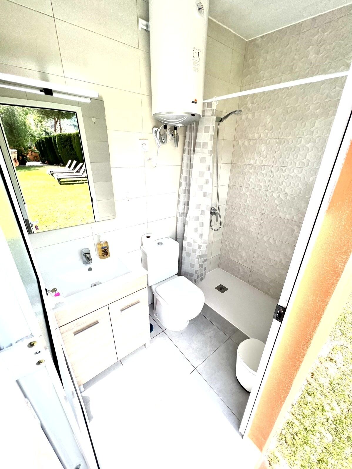 Een moderne badkamer met toilet, wastafel en douche. Boven de wastafel hangt een spiegel en door het raam is een tuin zichtbaar.