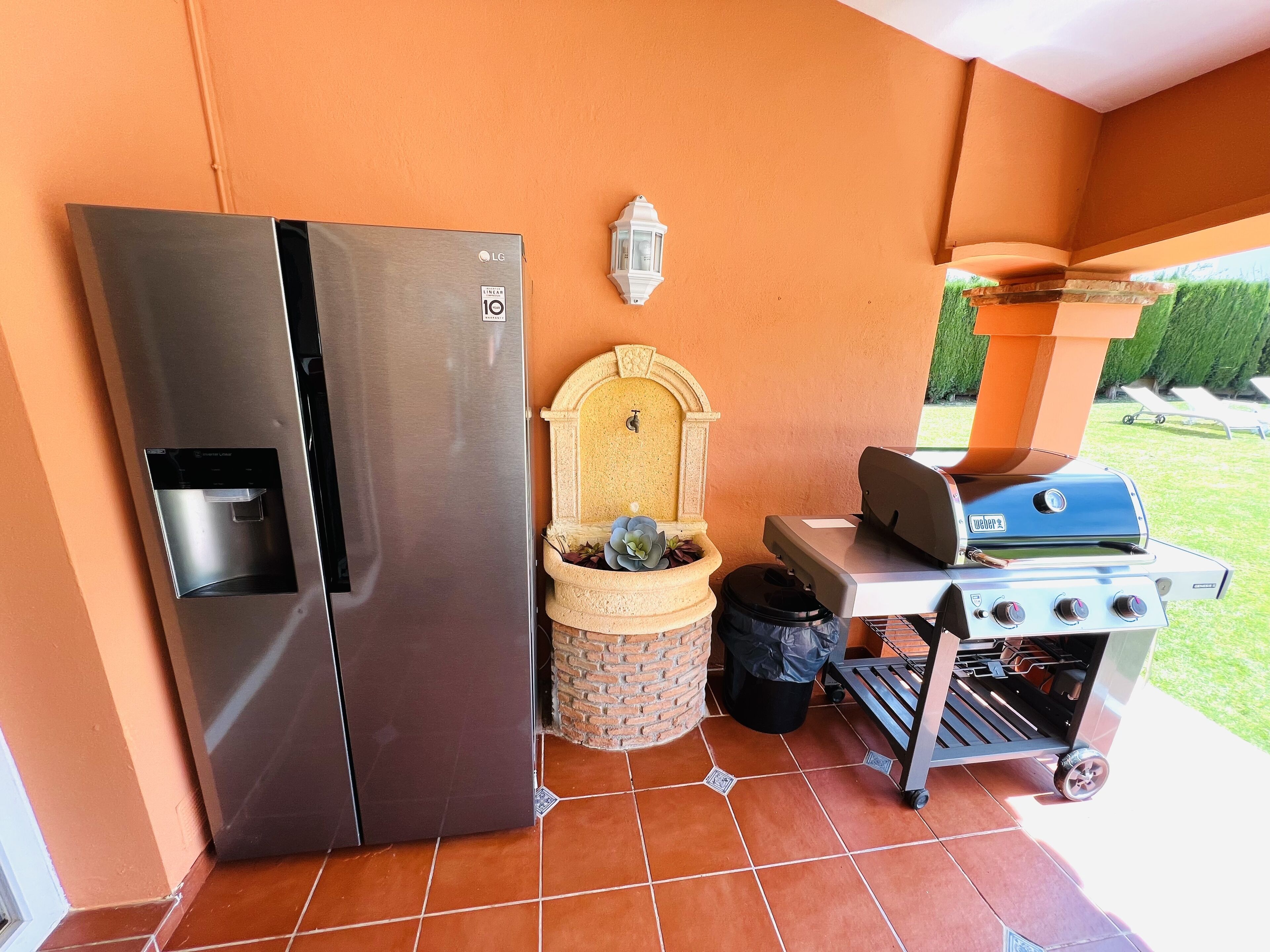 Un angolo cottura con frigorifero in acciaio inox, barbecue Weber, cestino e piccolo vaso con piante grasse.