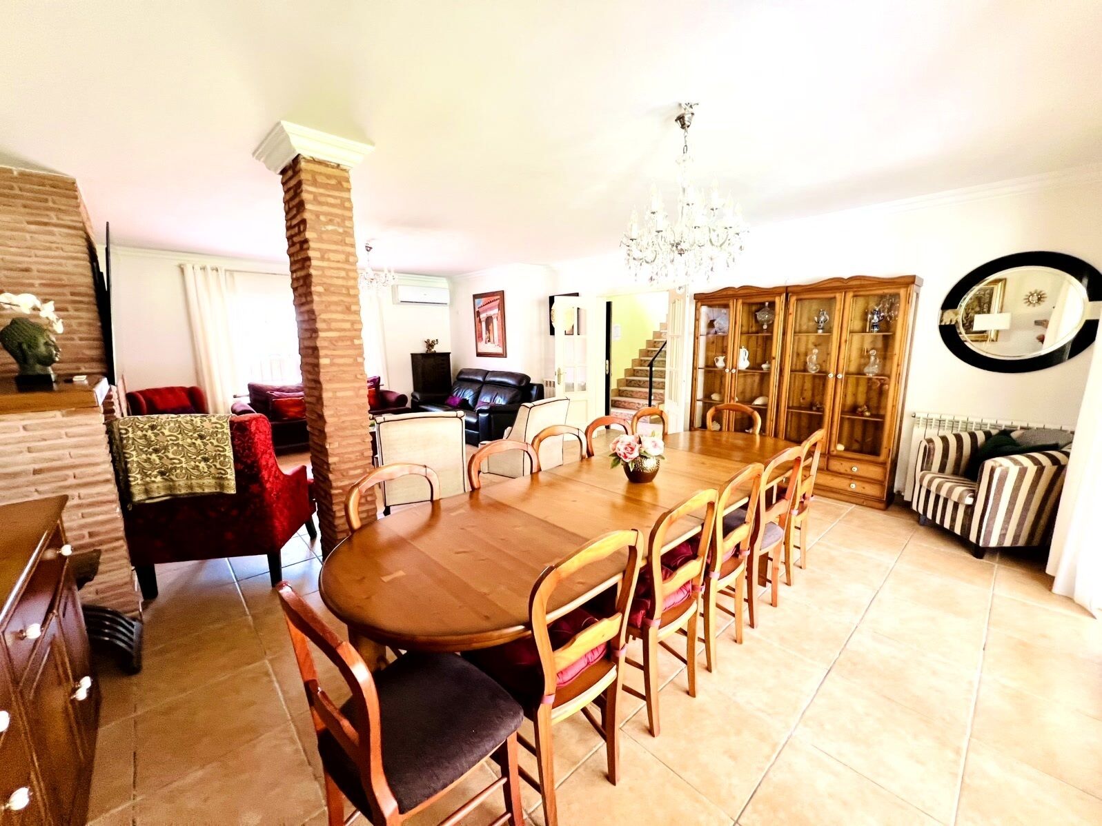 A dining area with a wooden table and chairs, a chandelier, a sofa, a cabinet, and a staircase leading to another room.