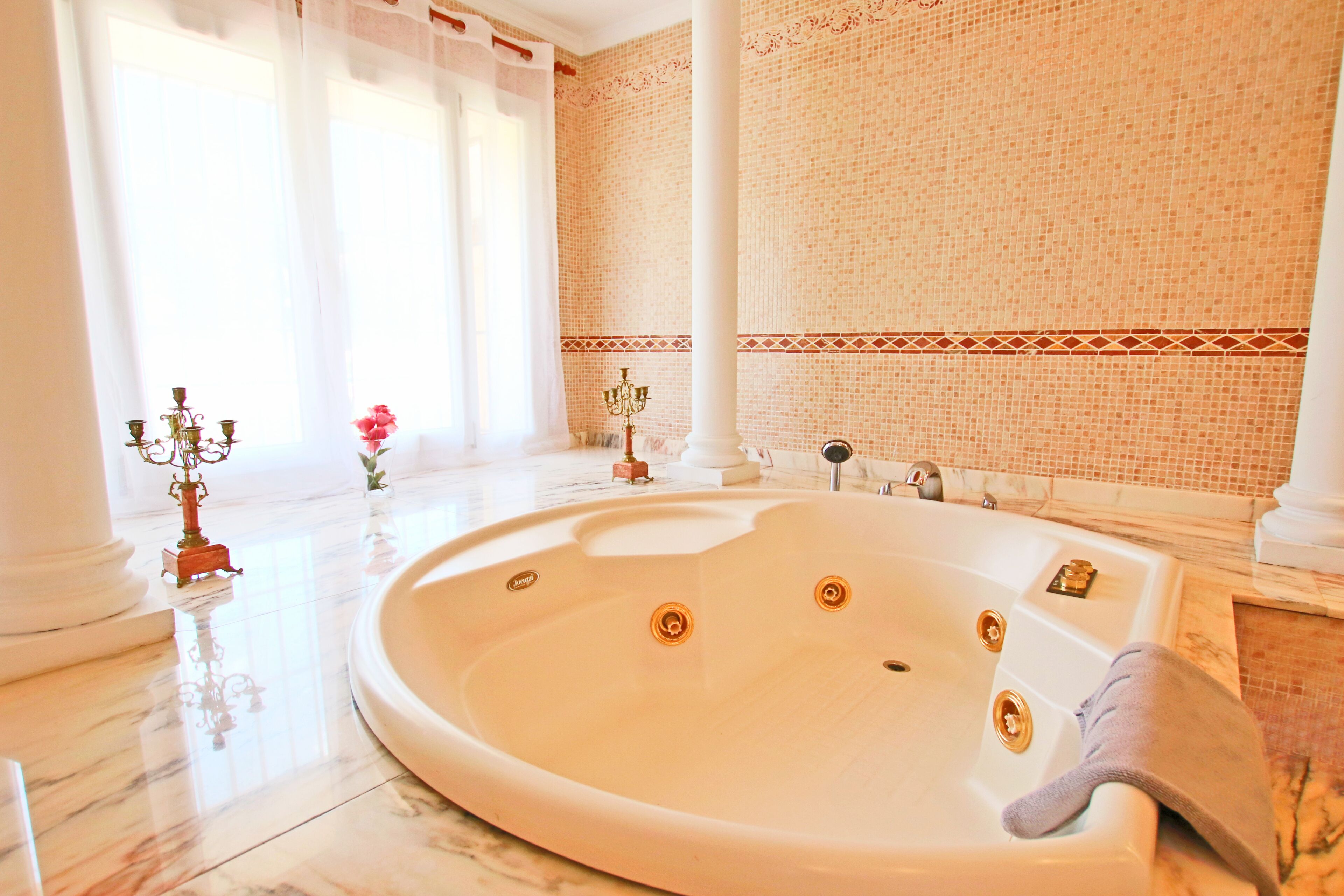 Une grande baignoire ronde et blanche, dotée de plusieurs robinets, est placée dans une pièce spacieuse dont le sol est carrelé et dont le mur est orné d’une bordure décorative.